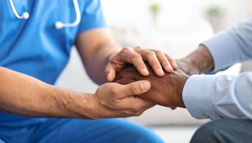 A doctor in blue scrubs holding the hands of an African-American man