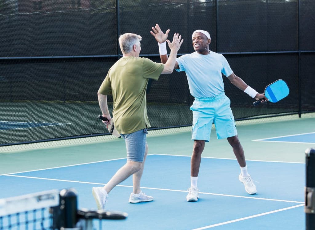 Two men playing pickleball and high fiving one another while their paddles are in their other hand