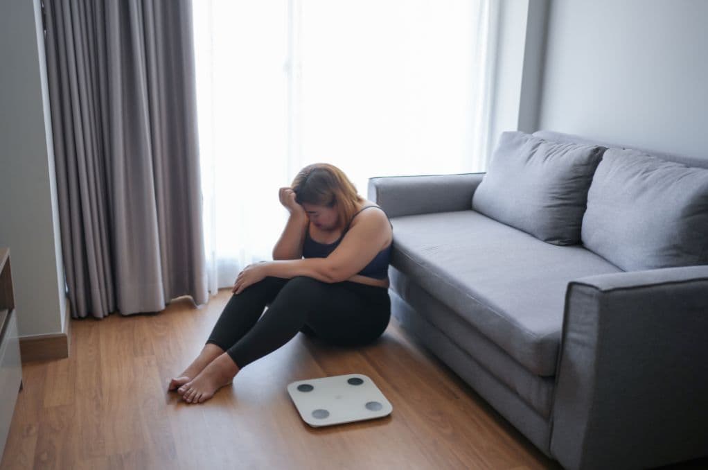 A woman sitting on the floor next to a body weight scale with her arms resting on her knees and her head in her hands looking upset
